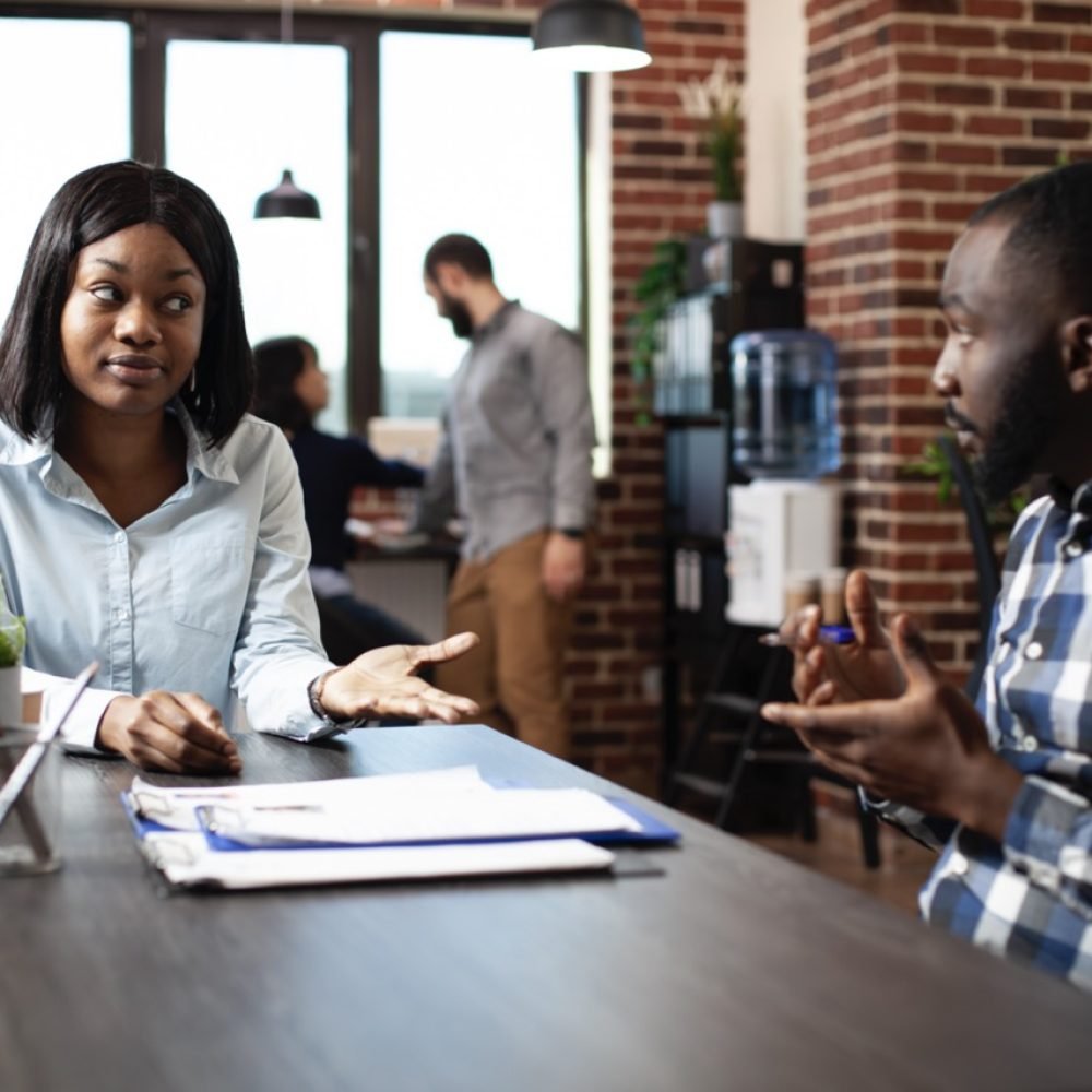 Young black woman explaining her work experiences and expectations to professional hiring manager. Male recruiter and female candidate engage in a formal discussion in a modern office.