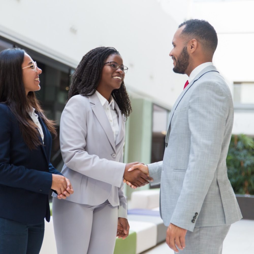 Happy joyful diverse business partners greeting each other in hallway. Business man and women standing, shaking hands with each other, talking and smiling. Successful partnership concept