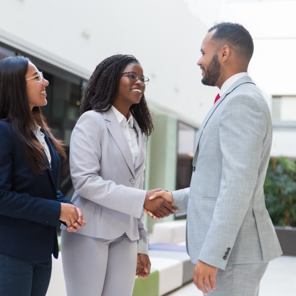Happy joyful diverse business partners greeting each other in hallway. Business man and women standing, shaking hands with each other, talking and smiling. Successful partnership concept